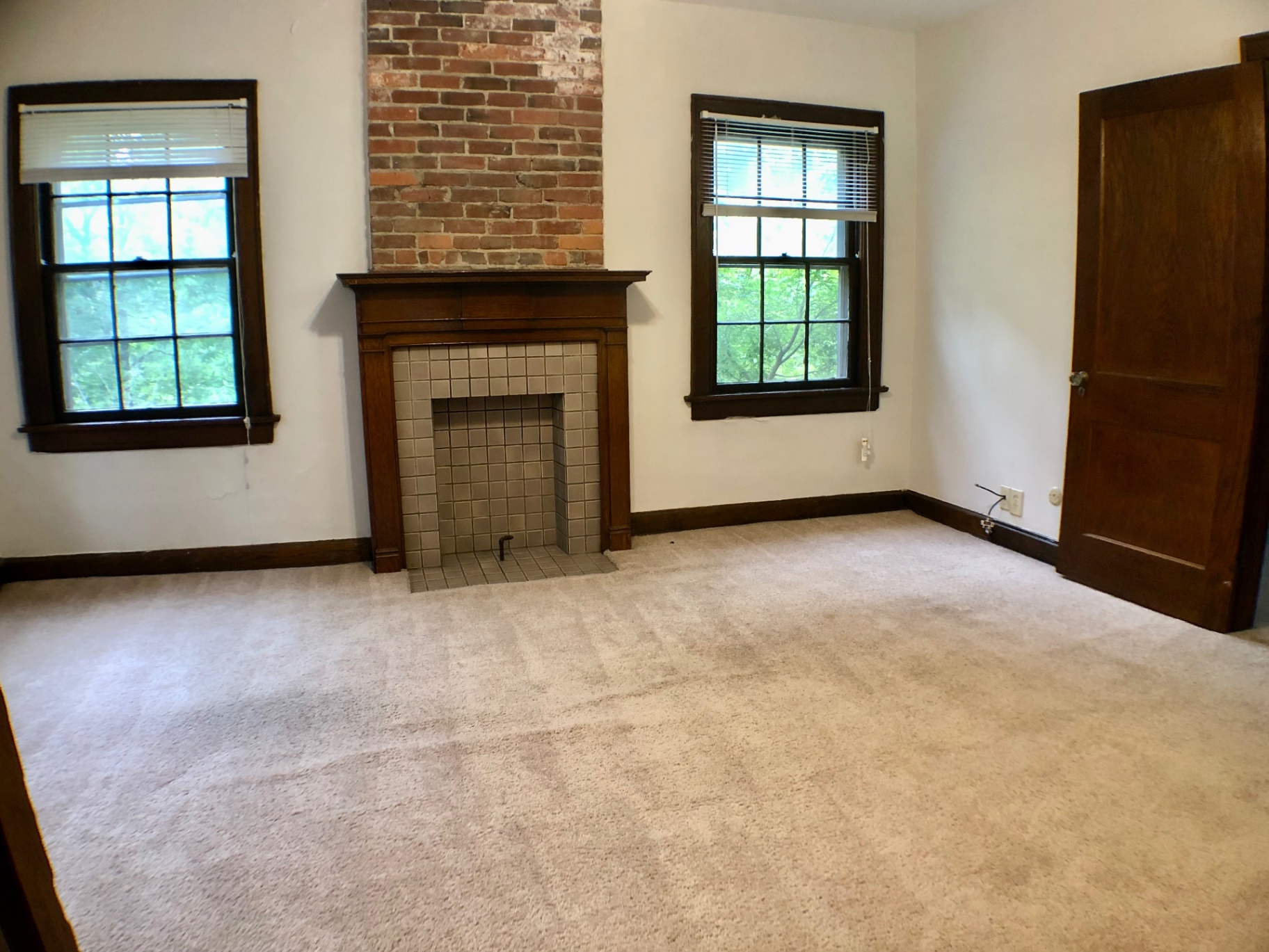 2054 Iuka Ave. D living room with original tile fireplace, wood mantel, exposed brick chimney, and dual windows overlooking the ravine
